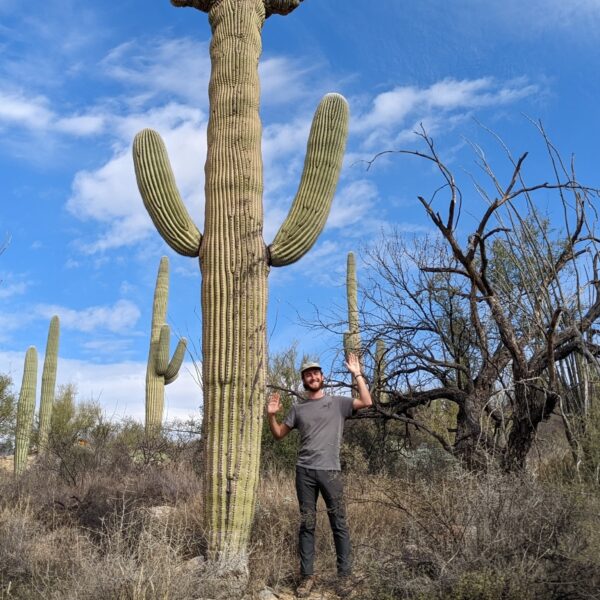 Lawton Bryant stands beside a tall cactus with arms raised in a desert landscape, under the vast blue sky.