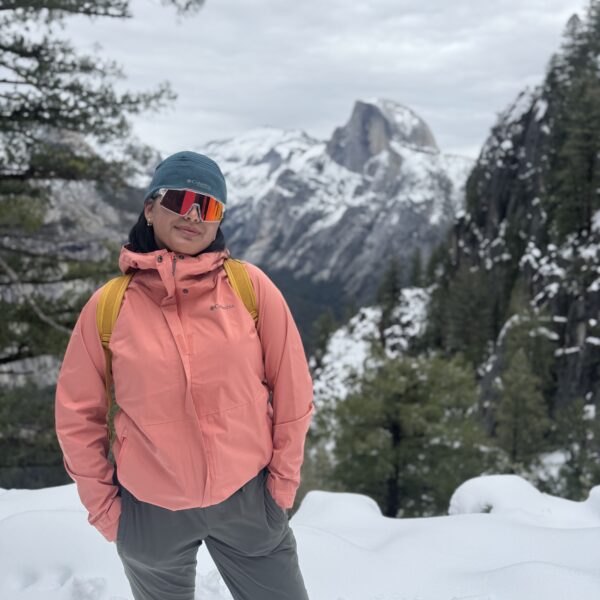 A person in a pink jacket and sunglasses stands on snowy ground with snowy trees and a mountain in the background.