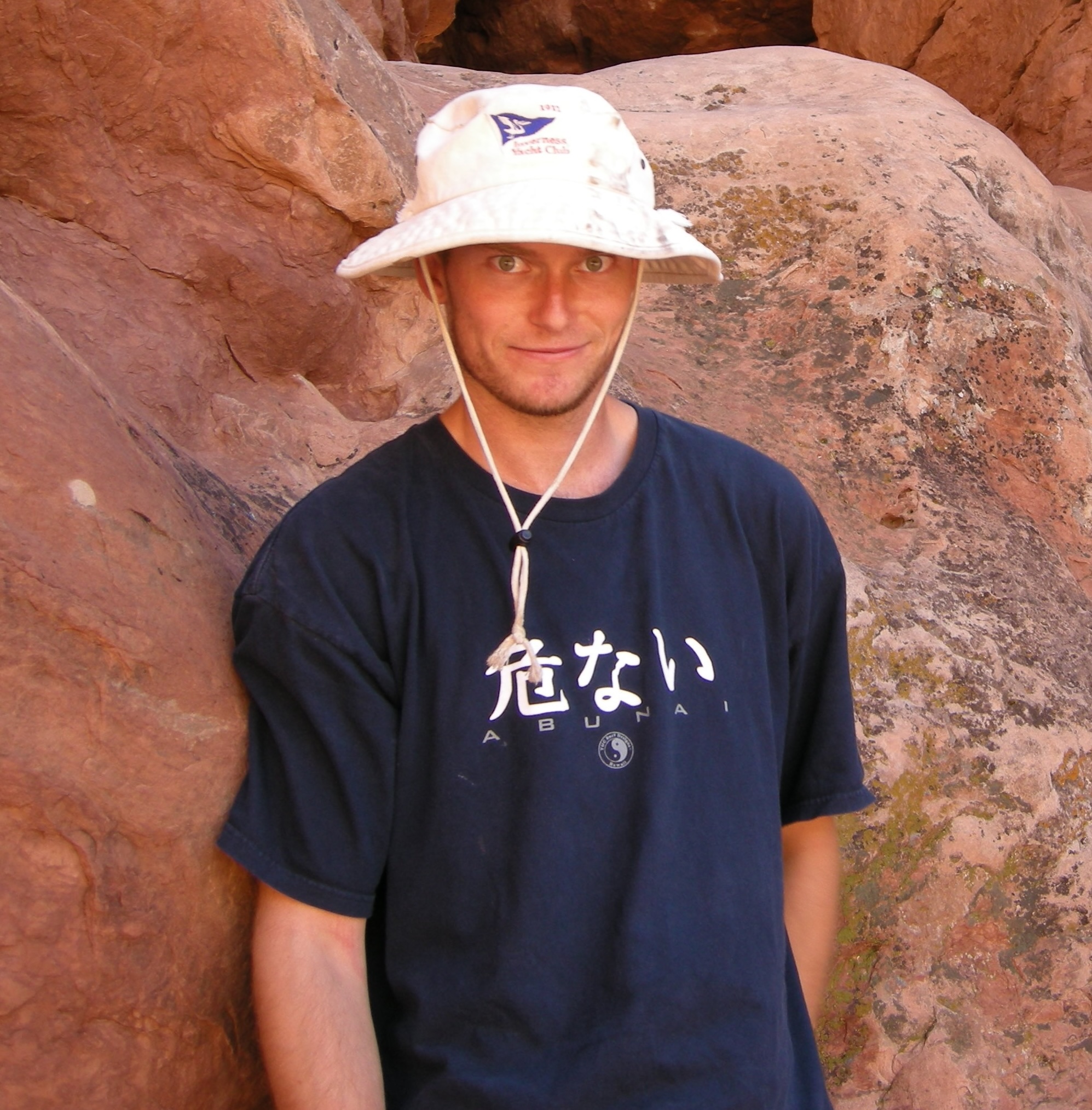 A person stands in front of rock formations wearing a white hat and a black T-shirt with Japanese characters and the word "ABUNAI.