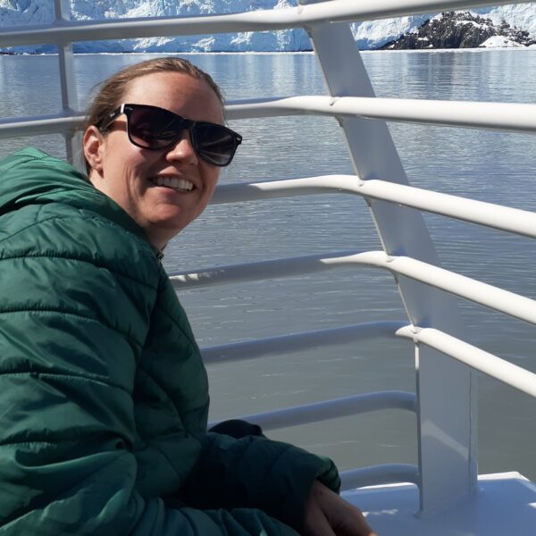 Laura Brooker, in a green jacket and sunglasses, smiles while sitting on a boat with icy water and glaciers in the background.