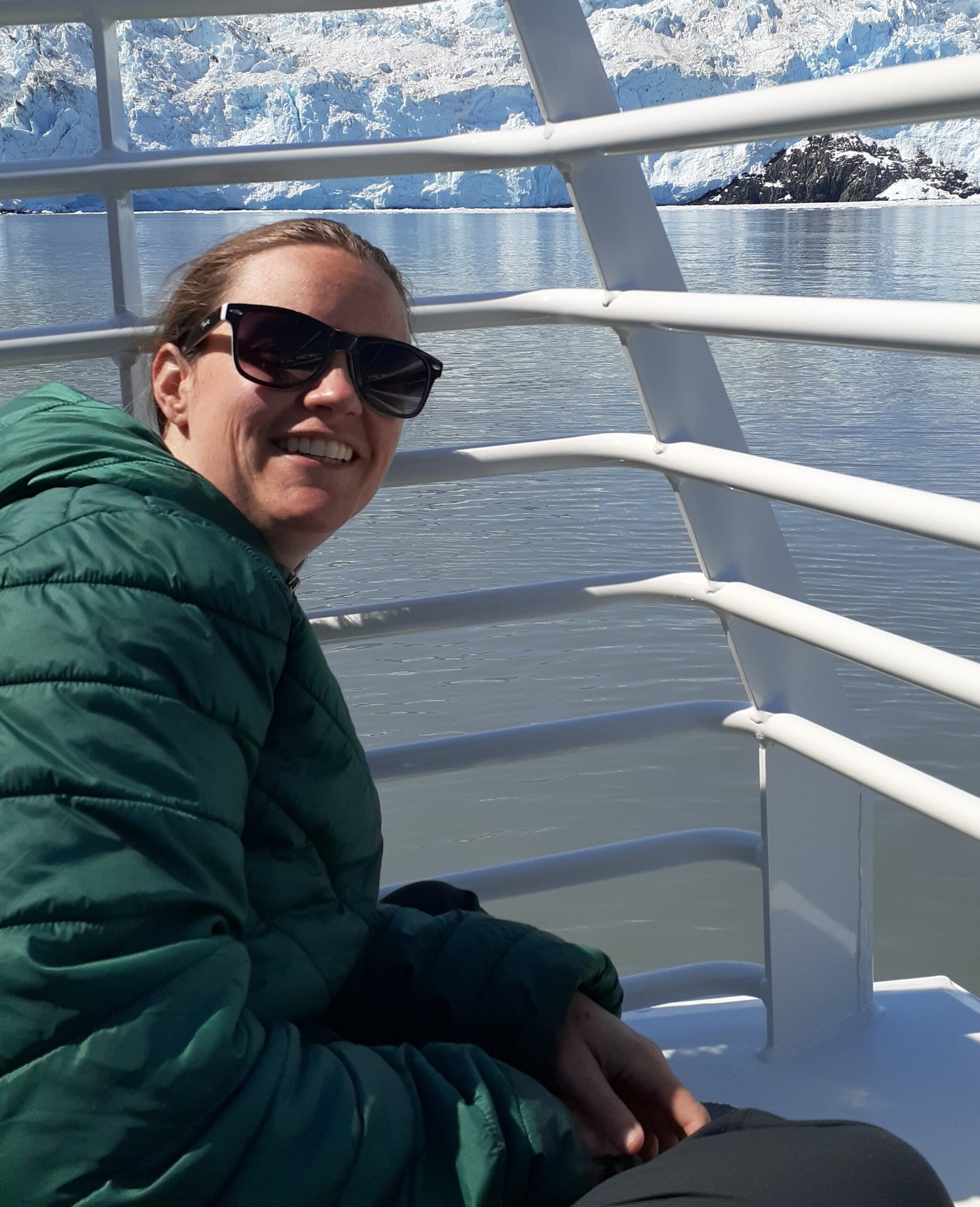 Laura Brooker, in a green jacket and sunglasses, smiles while sitting on a boat with icy water and glaciers in the background.