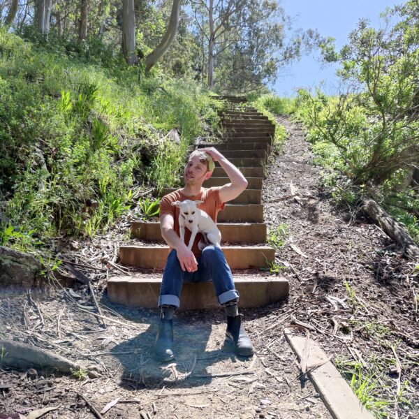 Elan Spitzberg is sitting on outdoor steps, surrounded by lush greenery, gently holding a white dog.