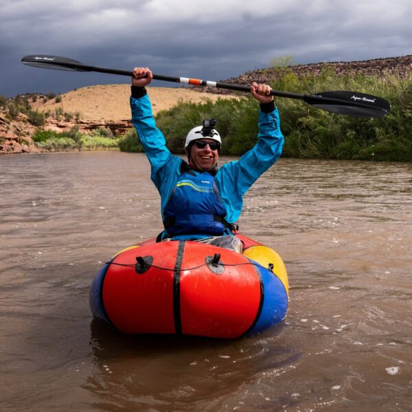 Person in a blue jacket and helmet holds a paddle overhead while sitting in a colorful inflatable kayak on a river, with a cloudy sky and desert landscape in the background.