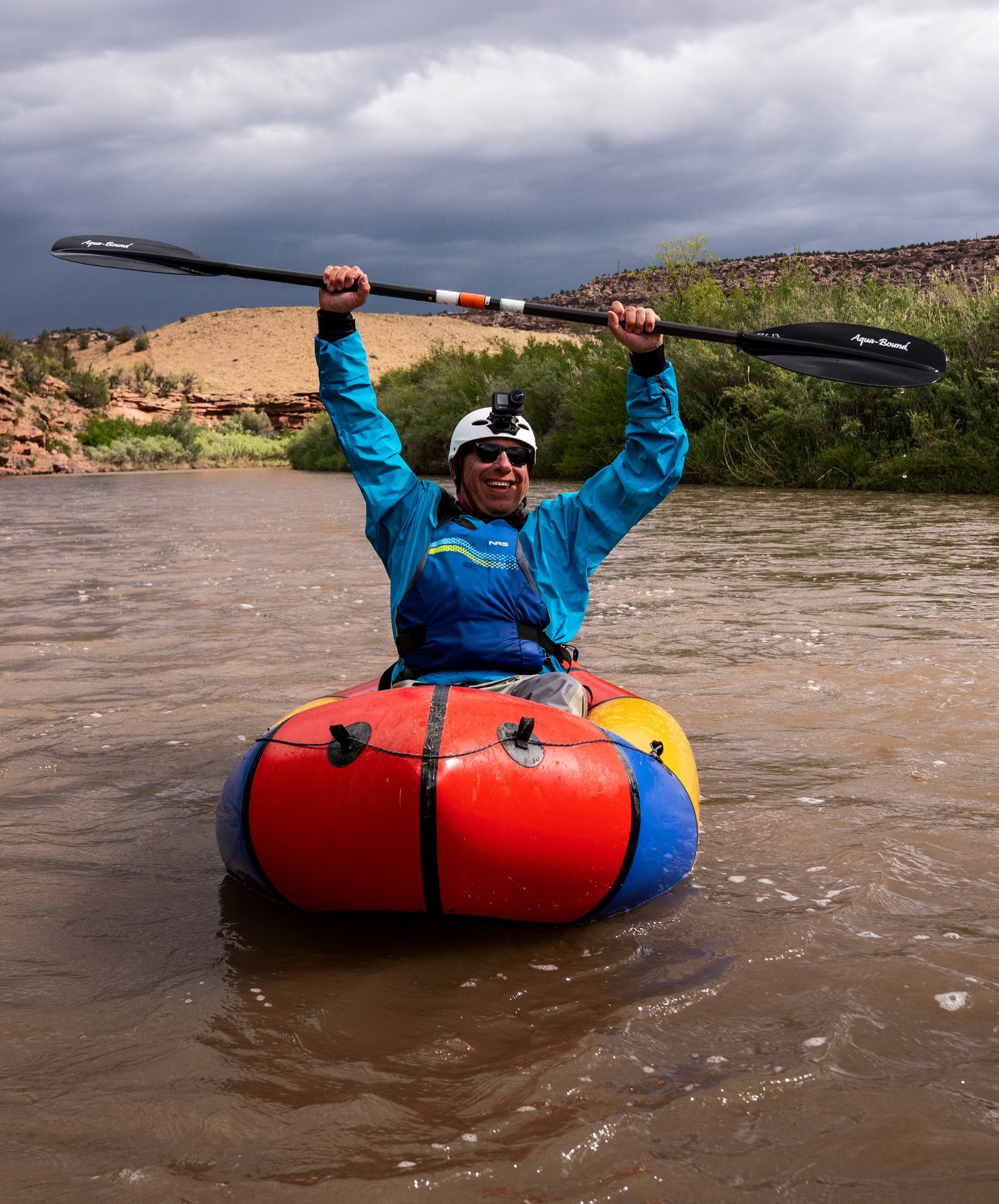 Person in a blue jacket and helmet holds a paddle overhead while sitting in a colorful inflatable kayak on a river, with a cloudy sky and desert landscape in the background.