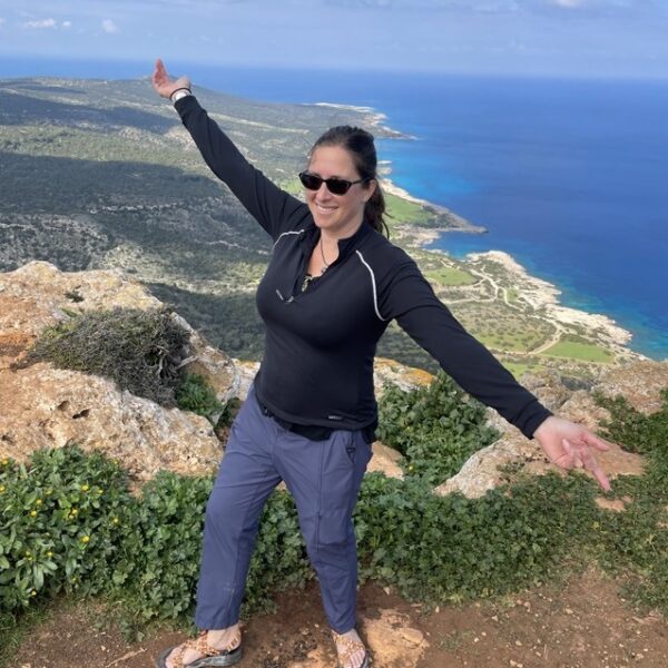 Jolie Ginsburg, wearing sunglasses and outdoor clothing, stands on a rocky hill with arms outstretched, overlooking a coastline and ocean under a partly cloudy sky.