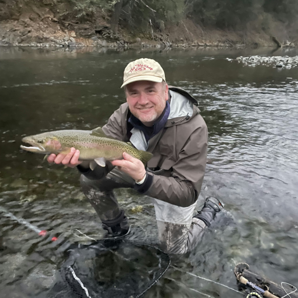 Brian Sugrue kneels in a shallow river, smiling and holding a large fish; a fishing rod and net rest on the riverbed beside him.