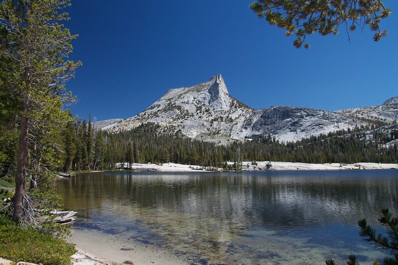 A mountain with a sharp peak rises above a forest and a clear, reflective lake, echoing the hidden gems of Yosemite’s valley floor beneath a bright blue sky.