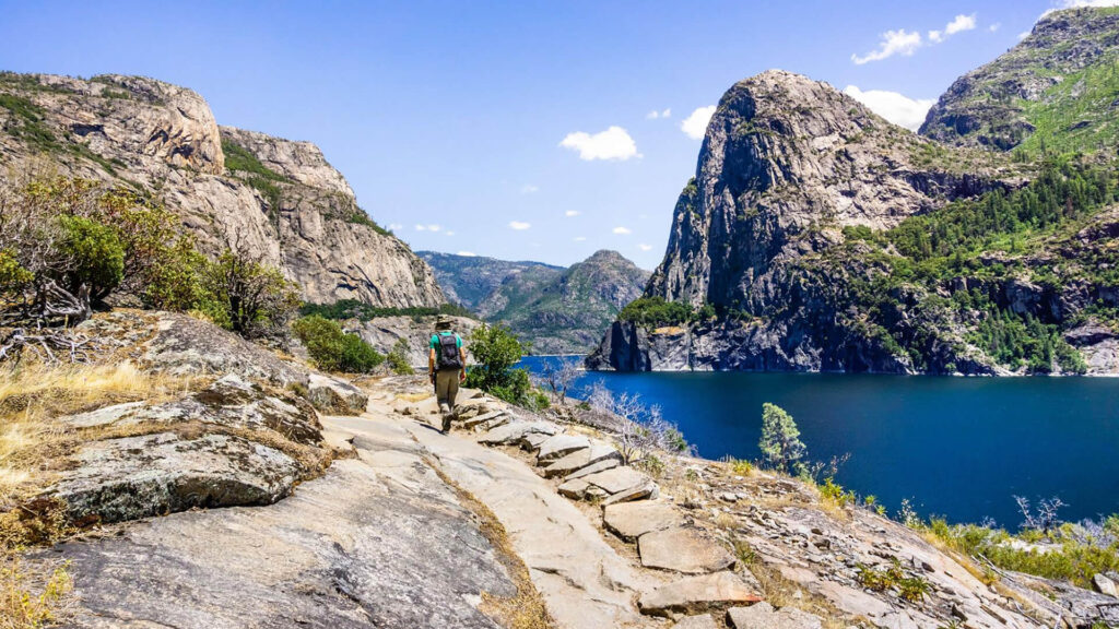 A hiker with a backpack walks along a rocky trail beside a blue lake in Yosemite, surrounded by steep mountains and clear skies—a hidden gem off the Valley Floor.