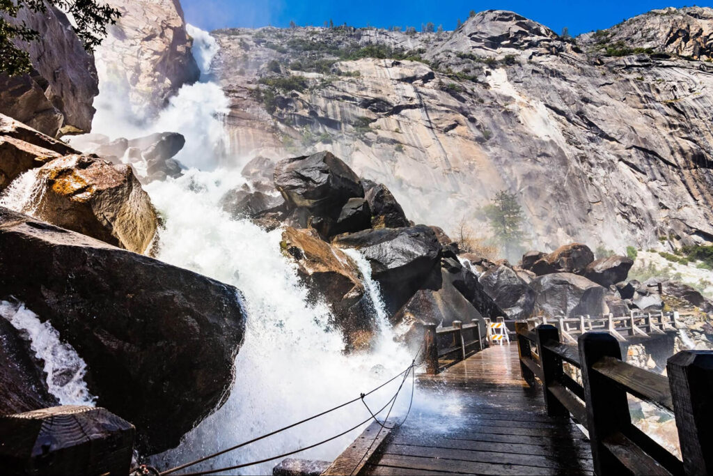 A powerful waterfall cascades down large rocks beside a wooden footbridge, with mist rising and rocky cliffs in the background—one of Yosemite’s hidden gems on the valley floor under a clear blue sky.