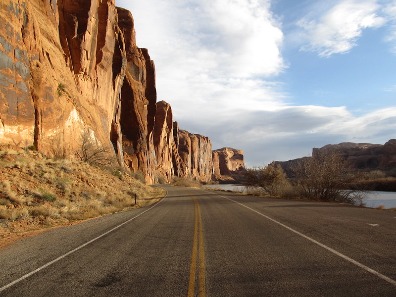 A paved road runs alongside red rock cliffs and a river under a partly cloudy sky, offering one of the iconic scenic drives of the American Southwest, with desert vegetation lining the roadside.