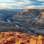 A wide view of the Grand Canyon with layered rock formations, a river winding through the canyon, and a rocky foreground under a partly cloudy sky.