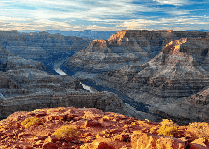 A wide view of the Grand Canyon with layered rock formations, a river winding through the canyon, and a rocky foreground under a partly cloudy sky.