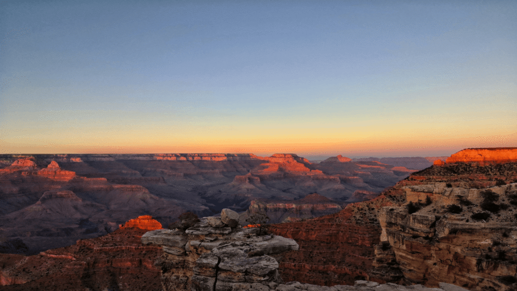 A view of the Grand Canyon at sunset, with rock formations and canyon layers illuminated by orange sunlight under a clear sky.