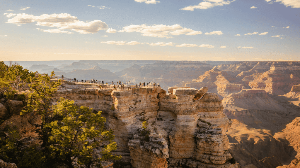 Tourists stand on a rocky overlook at the edge of the Grand Canyon, surrounded by layered cliffs and expansive canyon views under a partly cloudy sky.