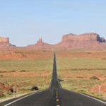 A straight road leads through a desert landscape toward red sandstone buttes and mesas under a clear blue sky.