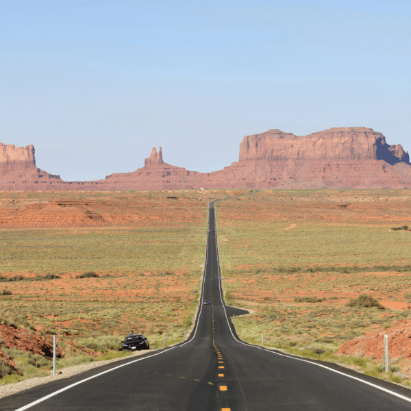 A straight road leads through a desert landscape toward red sandstone buttes and mesas under a clear blue sky.