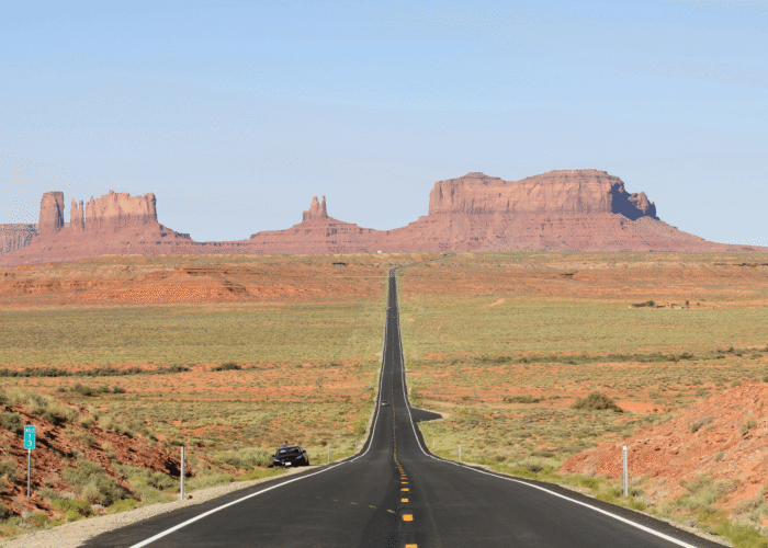 A straight road leads through a desert landscape toward red sandstone buttes and mesas under a clear blue sky.
