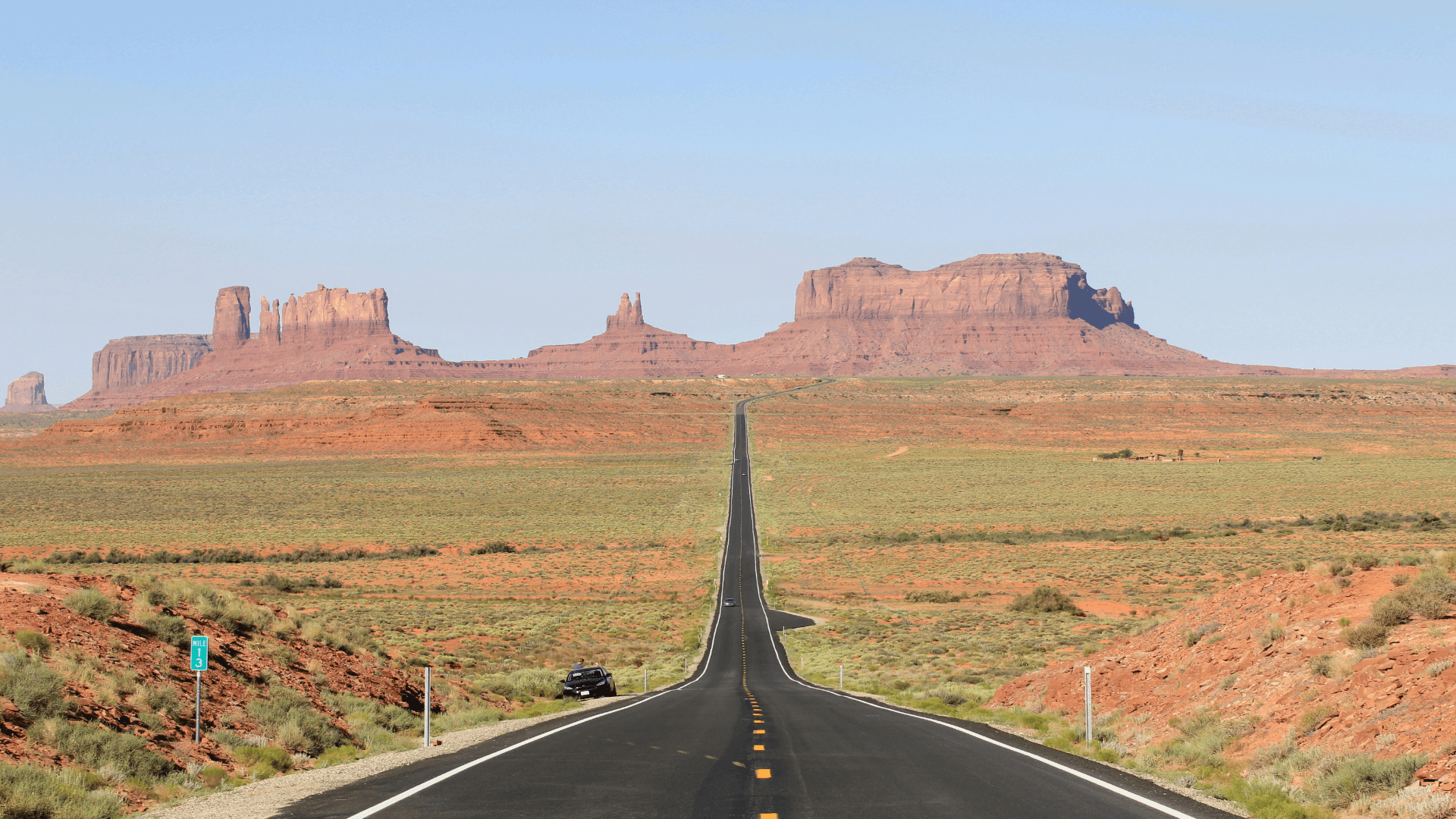 A straight road leads through a desert landscape toward red sandstone buttes and mesas under a clear blue sky.