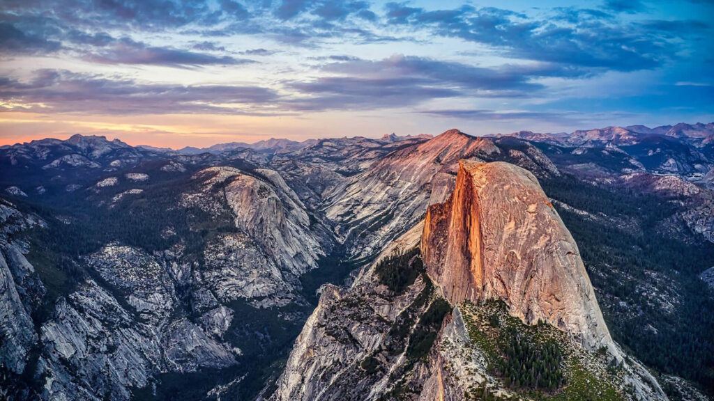 A panoramic view of Yosemite National Park at sunset, showcasing the best granite formation of Half Dome surrounded by rugged mountains and a partly cloudy sky—a true West Coast National Park travel style experience.