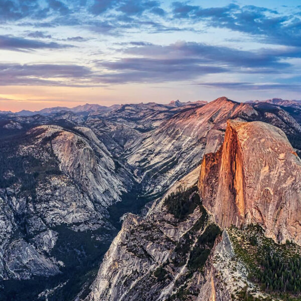 A panoramic view of Yosemite National Park at sunset, showcasing the best granite formation of Half Dome surrounded by rugged mountains and a partly cloudy sky—a true West Coast National Park travel style experience.