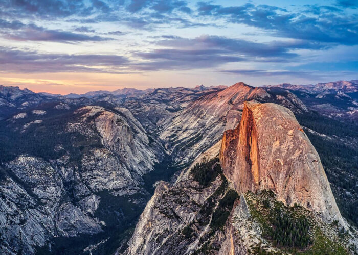 A panoramic view of Yosemite National Park at sunset, showcasing the best granite formation of Half Dome surrounded by rugged mountains and a partly cloudy sky—a true West Coast National Park travel style experience.