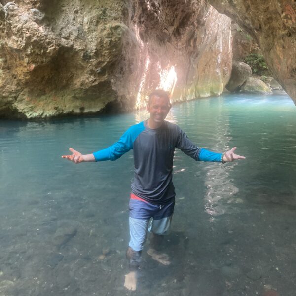Jonathan Yonts stands knee-deep in clear blue water inside a rocky cave, wearing a long-sleeve shirt and shorts, arms outstretched.