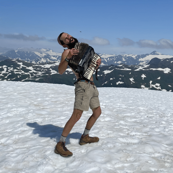 A man in shorts and boots plays an accordion enthusiastically on a snowy mountain slope with a backdrop of distant snow-capped peaks.