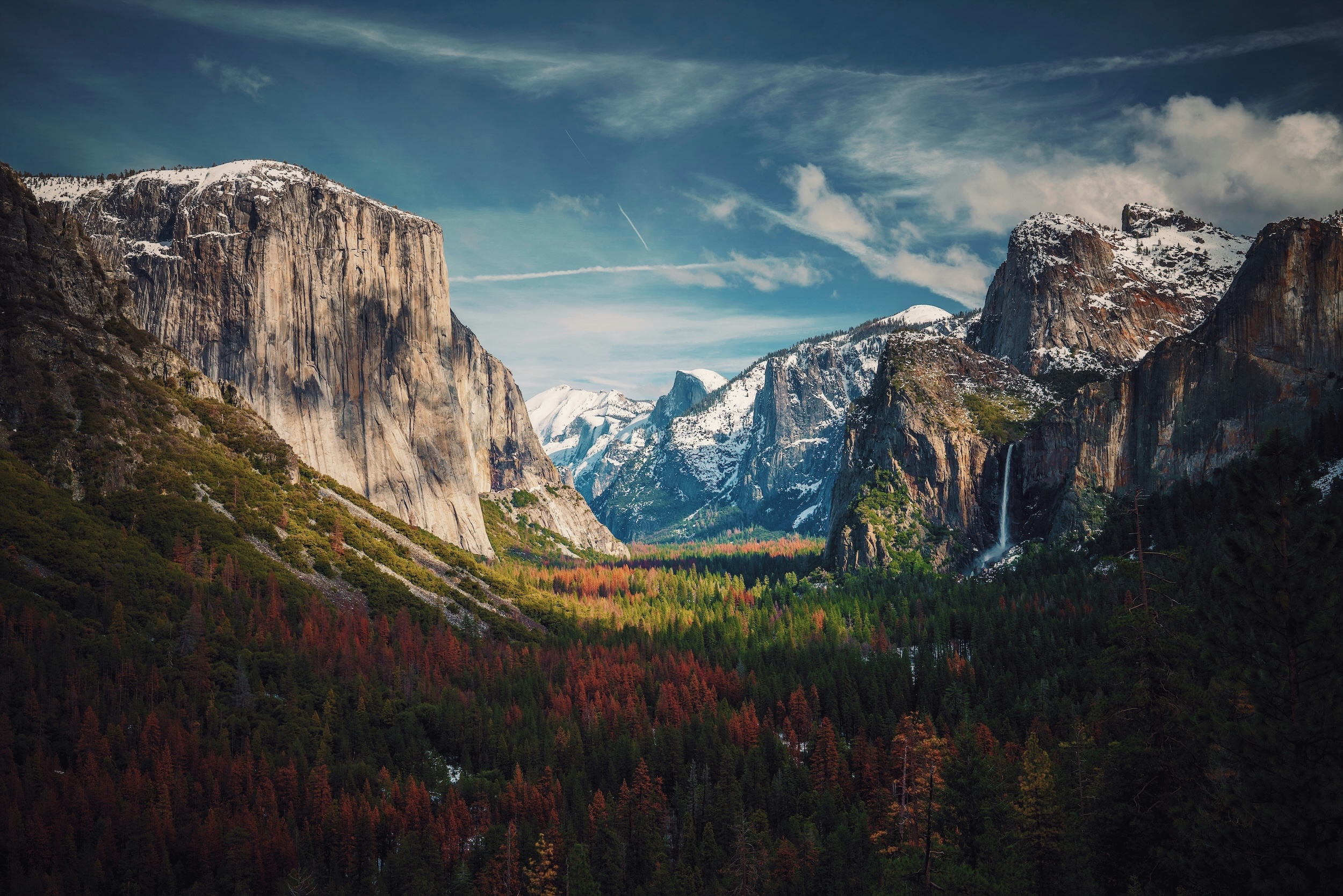 A panoramic view of Yosemite Valley with El Capitan on the left, Half Dome in the distance, and a forest of green and red trees below under a partly cloudy sky