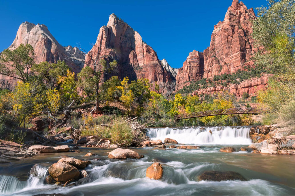 A river with small waterfalls flows over rocks, surrounded by trees and red rock cliffs under a clear blue sky. A wooden bridge spans the river in the background, evoking a travel style perfect for exploring a West Coast national park.