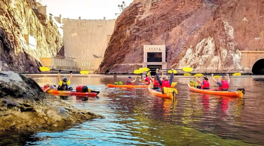 A group of people in orange kayaks paddle on a river near rocky cliffs and a large dam, enjoying a scenic Black Canyon kayaking tour just outside Las Vegas.