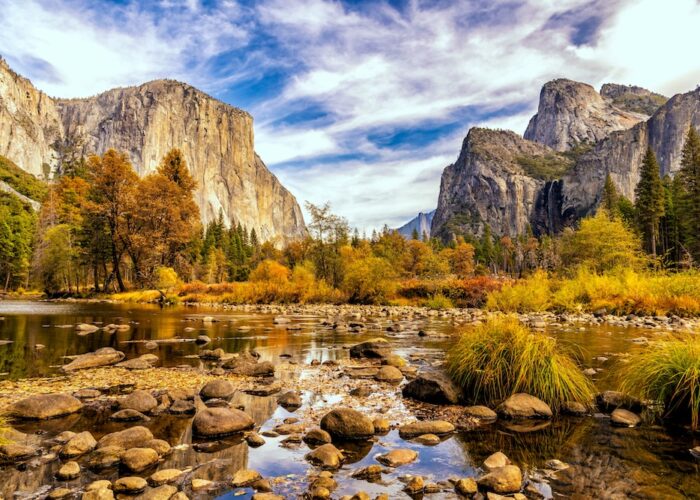 A river with rocks flows through a Yosemite valley surrounded by tall cliffs, pine trees, and autumn foliage—a magical escape under a partly cloudy November sky.