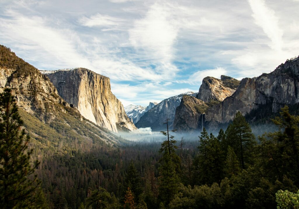 Experience a magical escape in November with this view of Yosemite Valley, featuring granite cliffs, pine trees, a misty valley floor, and a partly cloudy sky.