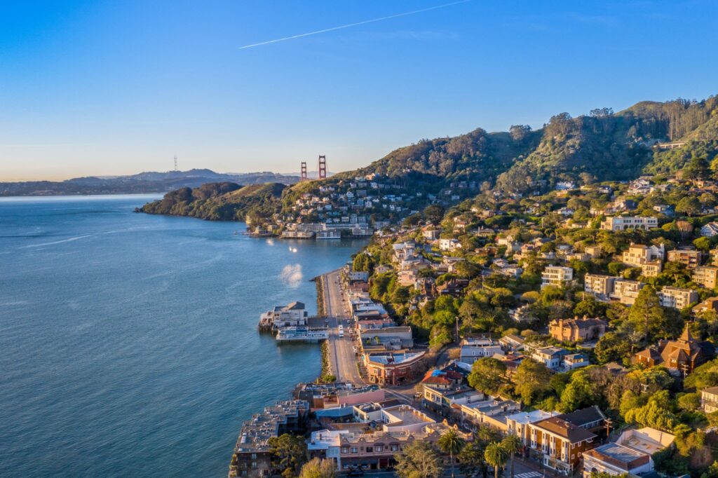 Aerial view of a Sausalito with hillside houses, a waterfront road, and the Golden Gate Bridge in the background under a clear blue sky—perfect for any San Francisco Neighborhood Guide.