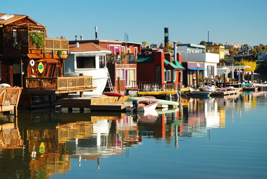 Colorful houseboats are docked side by side along a calm waterfront in this San Francisco Neighborhood Guide, with their reflections shimmering in the water under a clear blue sky.