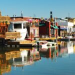 Colorful houseboats are docked side by side along a calm waterfront in this San Francisco Neighborhood Guide, with their reflections shimmering in the water under a clear blue sky.